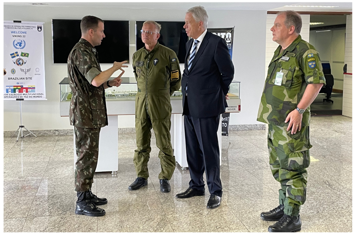 Quatro homens em pé. Três deles à direita escutam um à esquerda. Na ordem, da esquerda para a direita, dois militares, um civil e outro militar.